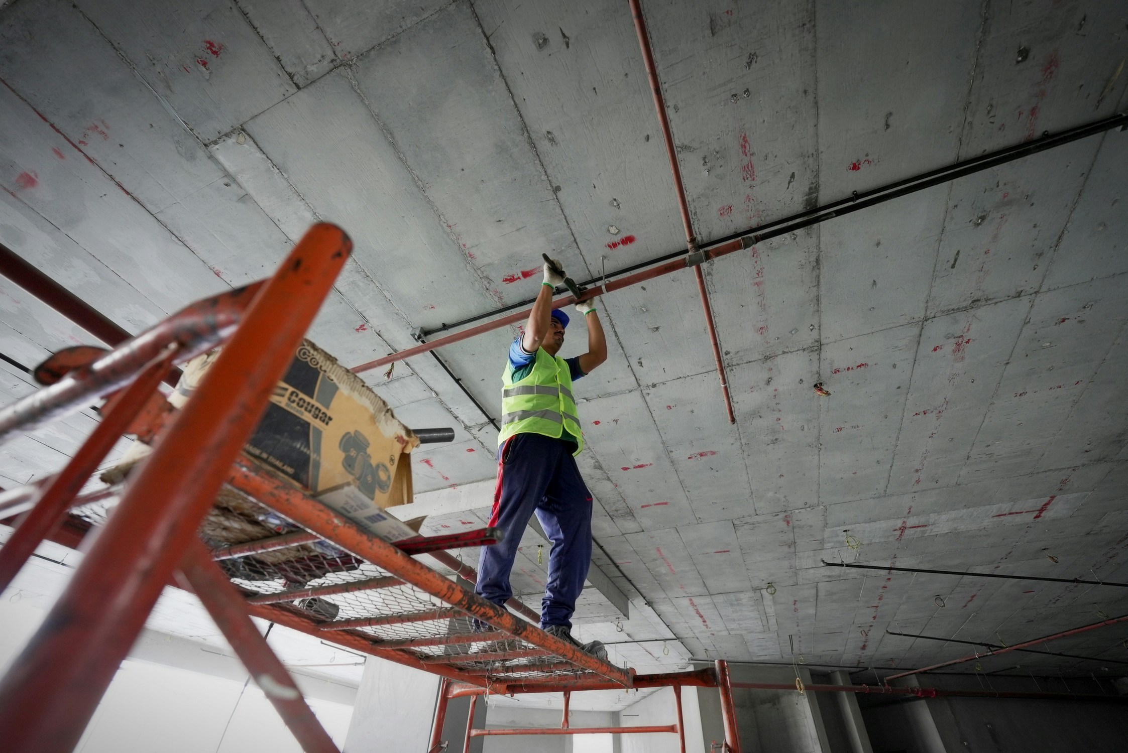 Un trabajador de la construcción con un chaleco de seguridad amarillo neón y guantes azules se encuentra de pie sobre un andamio y trabaja con tuberías unidas a un techo de hormigón. El lugar de trabajo está en un interior con un aspecto industrial e inacabado.