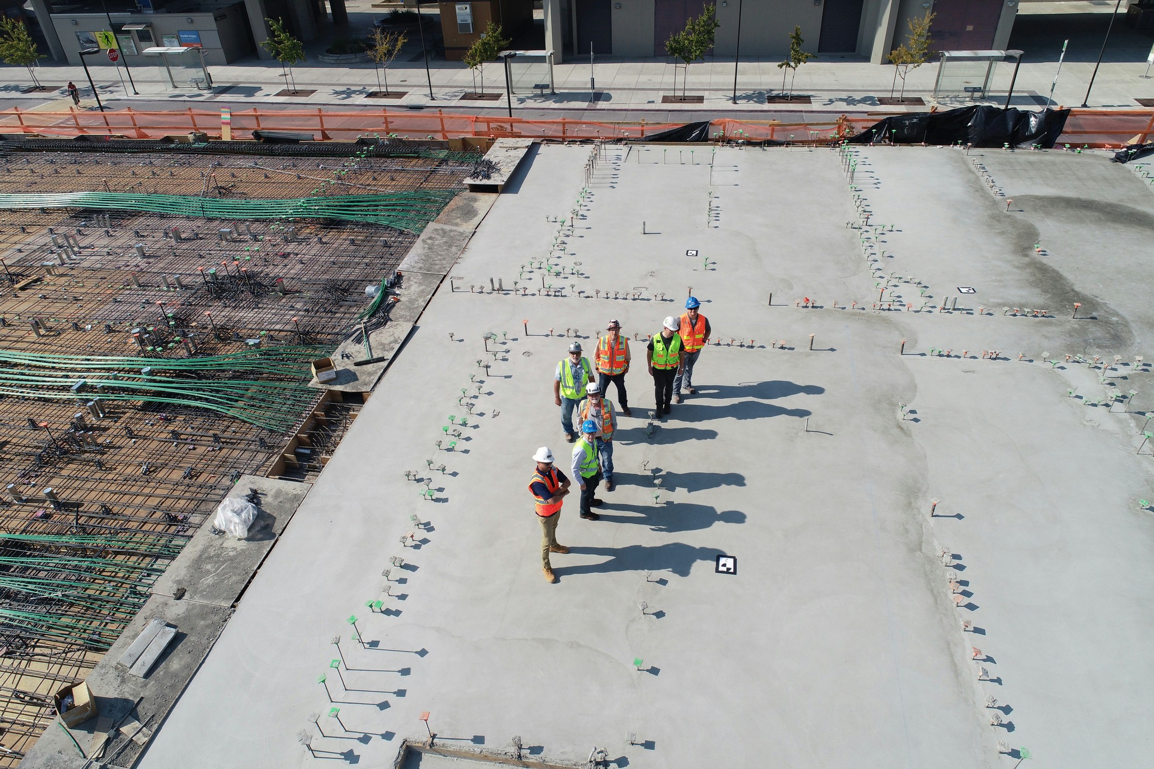 Vista aérea de siete trabajadores de la construcción con chalecos de seguridad y cascos, de pie sobre los cimientos de hormigón de un edificio parcialmente terminado. El lugar tiene barras de refuerzo, equipos y materiales expuestos esparcidos por todas partes. Parecen estar discutiendo el proyecto.